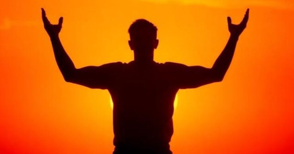 A person standing confidently against a bright sunset sky, arms slightly raised, powerful silhouette, vibrant orange and red tones, motivational atmosphere.